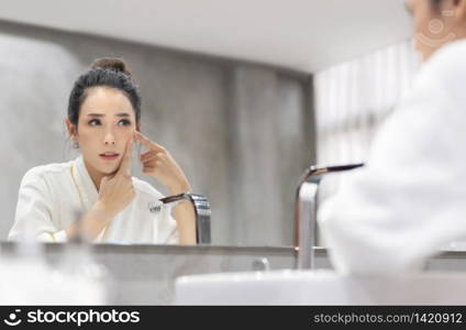 Stressed Asian woman in white Bathrobe looking at pimple on face into the mirror and try to squeeze it. Worried Young Beautiful girl try to remove her acne in bathroom. Skin care concept.