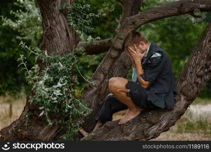 Stressed businessman in torn suit sitting on the tree on desert island. Business risk, collapse or bankruptcy concept