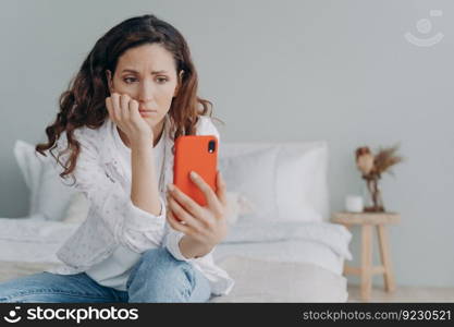 Stressed caucasian girl is reading morning news looking at smartphone display. Young woman is chatting on phone or browsing internet at home in her bedroom. Lady is doom scrolling.. Stressed caucasian woman is doom scrolling. Girl reading morning news on smartphone in her bedroom.