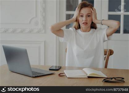 Stressed female holding head in hands, thinking of problem solving, looking at camera, sitting at desk with laptop. Worried young woman feeling anxious sits in workplace. Deadline, burnout concept.. Stressed female holding head in hands, thinking of problem solving, sitting at desk with laptop