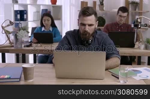 Stressed hipster man with beard working on laptop at desk in busy creative office and crumpling paper. Exhausted businessman under stress due to excessive work while colleagues working on background.