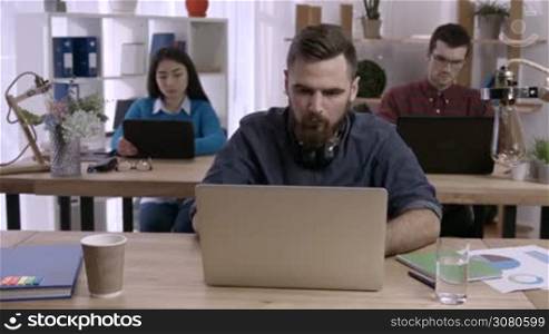 Stressed hipster man with beard working on laptop at desk in busy creative office and crumpling paper. Exhausted businessman under stress due to excessive work while colleagues working on background.