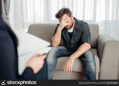 Stressed male patient at psychologist, professional psychology support. Female doctor writes notes in notepad, man sitting on sofa. Sad patient at psychologist, psychology support