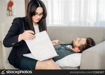Stressed man lies on sofa at psychotherapist reception. Female doctor writes notes in notepad, professional psychology support. Man lies on sofa at psychotherapist reception