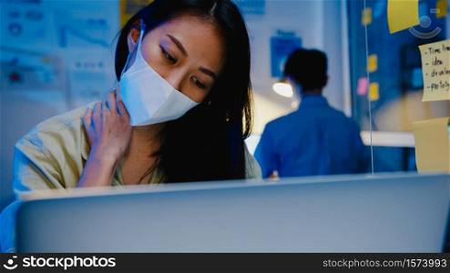 Stressed tired young Asia woman wear face mask using laptop hard work having office syndrome , neck pain, while working overtime at office. Working from home overload at night, social distancing.