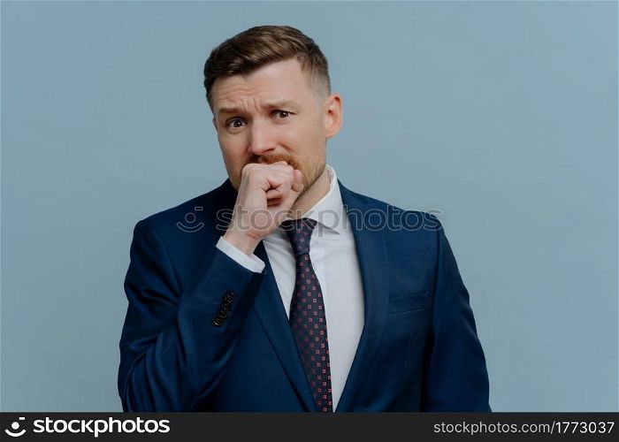 Stressed unshaved businessman in suit feeling nervous, depressed male office worker biting fist emotionally while standing against light steel blue background. Business problems and failure concept. Nervous businessman biting fist emotionally and looking at camera