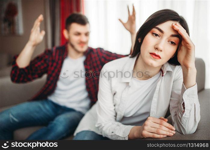 Stressed wife sits on couch, angered husband yells on her, family conflict. Unhappy man and woman in quarrel