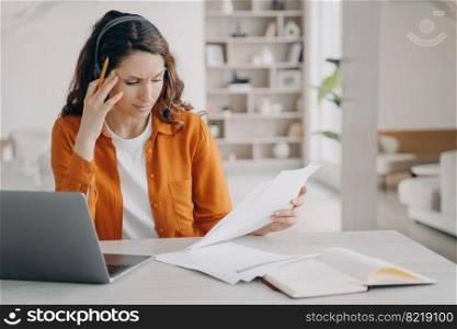 Stressed woman in headset looking through paper document, works at laptop at home. Female solves working problem. Pensive girl student stack with hard task, learning online. Remote job, education.. Female employee in headset looking through paper document, working at laptop at home. Remote work