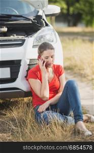 Stressed woman sitting at the broken car on roadside and talking by phone