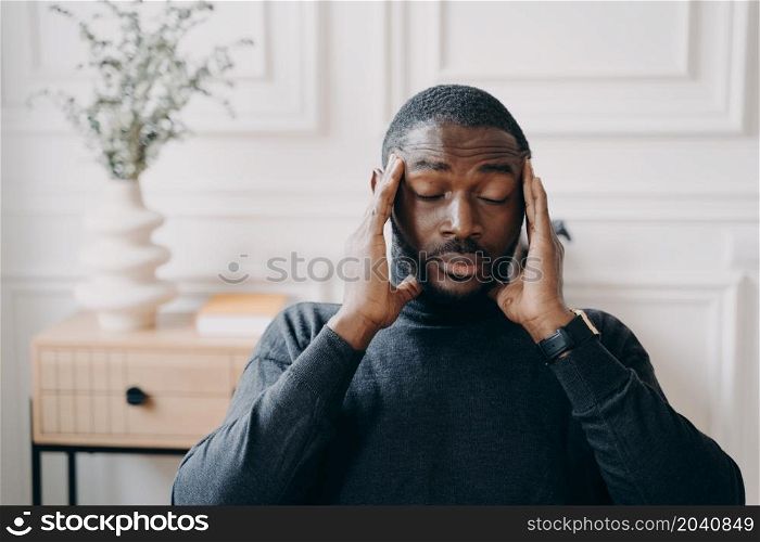 Stressed young african man employee with frustrated face expression suffering from headache, sitting with closed eyes and hands on temples, worried american male office worker having problems at work. Stressed young african man employee with frustrated face expression suffering from headache