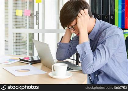 Stressed young Asian businessman sitting at desk looking at labtop and touching his temples with migraine headache of business problem or job failture in meeting room. Business stress and failure concept