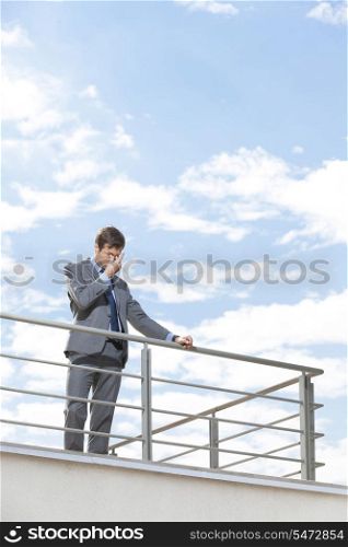 Stressed young businessman rubbing eyes on terrace against sky