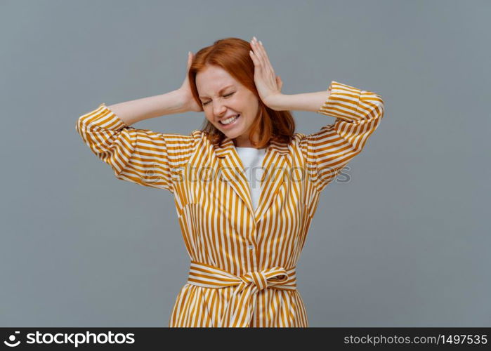 Stressful redhead woman feels pressure and terrible headache, grabs head, clenches teeth from pain, wears yellow striped robe, stands against grey background. Housewife suffers from migraine