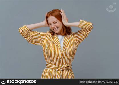 Stressful redhead woman feels pressure and terrible headache, grabs head, clenches teeth from pain, wears yellow striped robe, stands against grey background. Housewife suffers from migraine