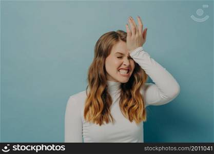 Stressful woman with brown hair wearing casual clothes holding hand on head having disappointment and stressful situation, looking frustrated with eyes closed isolated on blue with free space. Stressful woman holding hand on head having stressful situation, looking frustrated with eyes closed
