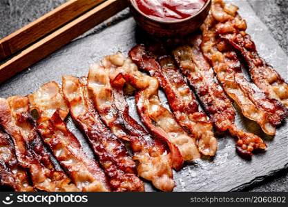 Strips of fried bacon on a stone board with tomato sauce. On a black background. High quality photo. Strips of fried bacon on a stone board with tomato sauce.