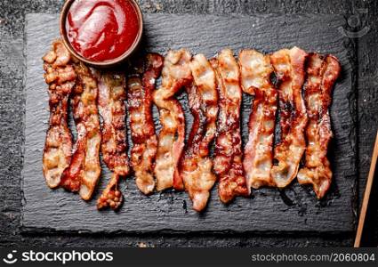 Strips of fried bacon on a stone board with tomato sauce. On a black background. High quality photo. Strips of fried bacon on a stone board with tomato sauce.