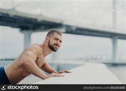 Strong bearded Caucasian man trains muscles and does push ups exercises, tries to reach fitness goals, demonstrates his physical strength, has naked torso, poses outdoor over bridge background.