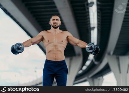 Strong bearded young man with muscular body raises arms with heavy barbells uses sport equipment dressed in sportswear trains actively outdoor. Healthy lifestyle workout and bodybuilding concept