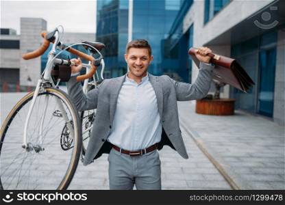 Strong businessman holds bicycle and briefcase at the glass office building in downtown. Business person riding on eco transport on city street, urban style