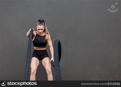 Strong female athlete in sportswear with ponytail looking at camera with frown and exercising with heavy battle ropes against black wall in gym. Aggressive sportswoman doing exercise with battle ropes
