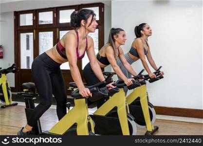 Strong female athletes in activewear smiling and looking away while exercising on bikes during cardio workout in daytime in gym. Sportswomen exercising on bicycles together