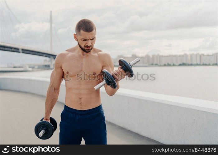 Strong fitness sporty bearded man with thick bristle does exercises with dumbbells, pumps up biceps, poses outdoor, has naked torso, breathes fresh air. Fitness, sport and lifestyle concept.