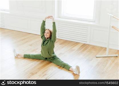 Strong flexible curly red haired woman does side leg split, raises hands, wants to have perfect body, wears sportsclothes, has training in ballet studio. Talented dancer has rehearsal indoor