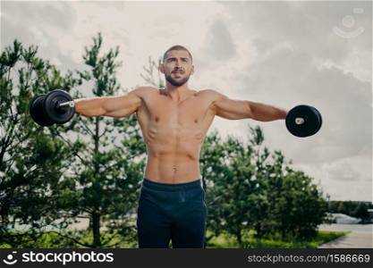 Strong handsome muscular bodybuilder pumps up arm muscles with barbells, does bidybuilding biceps exercises outdoor, poses with naked torso against green trees and sky, makes weight exercise