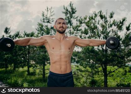 Strong healthy man stretches arms, makes weightlifting and exercises biceps with barbells, poses with naked muscular torso outdoor. Athletic shirtless motivated sportsman has workout in open air