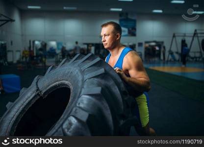 Strong male athlete doing strength exercise with truck tyre in gym, crossfit workout. Cross fit training in fitness club. Athlete doing exercise with truck tyre, crossfit