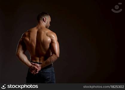 Strong male athlete poses in studio, back view, dark background. One man with athletic build, shirtless sportsman in jeans pants, active healthy lifestyle. Strong male athlete poses in studio, back view