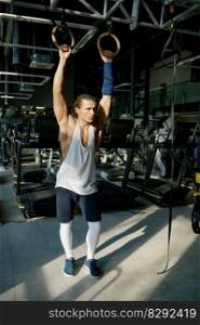 Strong male athlete with muscular perfect body standing with gymnastic rings in gym. Fit man taking break to rest and catch breath between sets. Strong male athlete with muscular body standing with gymnastic rings in gym