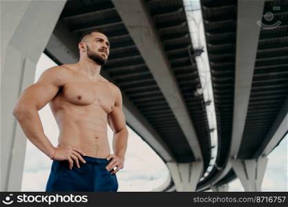 Strong man with naked torso keeps hands on waist, has muscular body after long training, focused into distance, stands under bridge, thinks about how to be healthy and fit. Sporty man outdoor