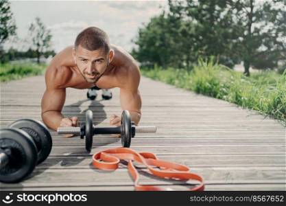 Strong motivated handsome man stands in plank pose, makes abdominal exercises, poses near sport equipment, listens music in wireless earphones, has workout outdoor, looks with serious expression