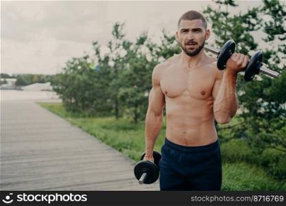 Strong muscular bodybuilder prepares for athletic competition, raises heavy dumbbells, trains outdoor, demonstrates his sport success, has naked torso and perfect body. Healthy lifestyle concept