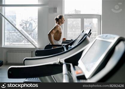 Strong muscular man running on treadmill at modern sport gym. Athletic male bodybuilder jogging on stationary machine doing cardio training. Side view. Strong muscular man running on treadmill at modern sport gym