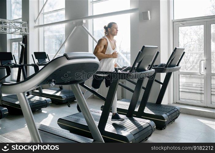 Strong muscular man running on treadmill at modern sport gym. Athletic male bodybuilder jogging on stationary machine doing cardio training. Strong muscular man running on treadmill at modern sport gym