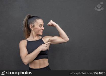 Strong sportswoman with ponytail smiling and pointing at muscle of bent arm while standing against black wall in gym. Muscular female athlete pointing at bicep
