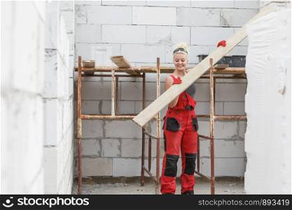 Strong woman in dungarees carrying big wooden plank board on her home construction site, building new home.. Woman carrying plank boards on construction site