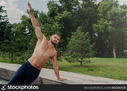 Strong young man stands in side plank on one arm, finds balance, poses at park near trees, practices yoga outdoor, leads active healthy lifestyle, has muscualr strong body. Motivated bodybuilder