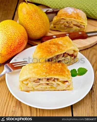 Strudel with pears in a white plate with a fork, pear, knife, napkin on the background of wooden boards