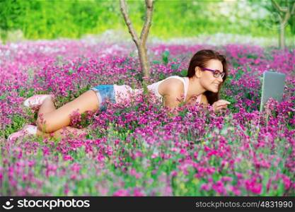 Student girl in the park, lying down on beautiful fresh flowers field and working on laptop, chatting in internet, learning in higher education