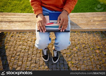 Student holding personal studying accessories as textbook, copy-book with pen and laptop while sitting on bench in university or college park. Student holding personal studying accessories high angle closeup view