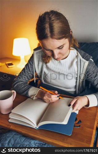 Student learning at home. Young woman making notes, reading and learning from notepad. Girl writing journal sitting in bed at home during quarantine