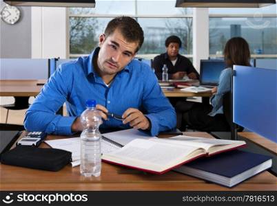 Student, looking into the camera, studying for his exams in a public library, with two other students in the background