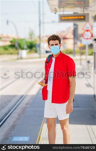 Student male wearing a surgical mask while waiting for a train at an outside station. Young man wearing a surgical mask while waiting for a train at an outside station