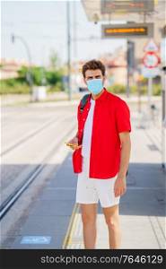 Student male wearing a surgical mask while waiting for a train at an outside station. Young man wearing a surgical mask while waiting for a train at an outside station