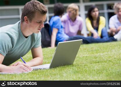 Student outdoors on lawn using laptop with other students in background (selective focus)