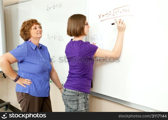 Student works algebra problem on the blackboard while her teacher looks on.